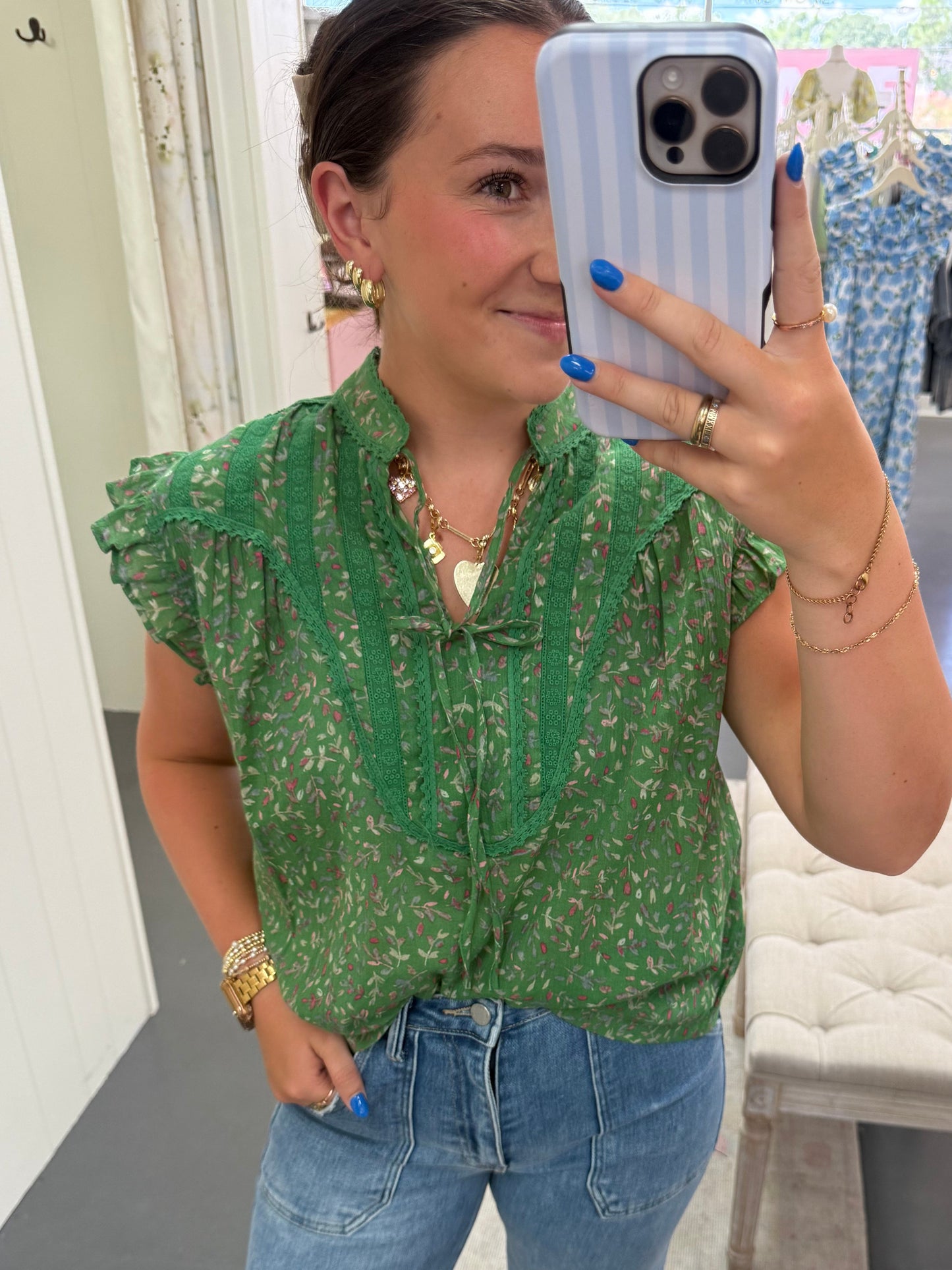 Person wearing a green floral top and blue jeans taking a mirror selfie.
