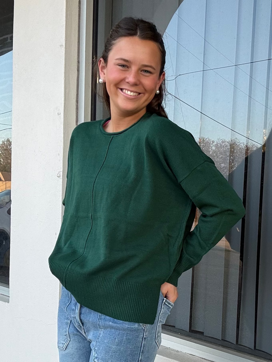 Woman wearing a green sweater and blue jeans standing in front of a window.