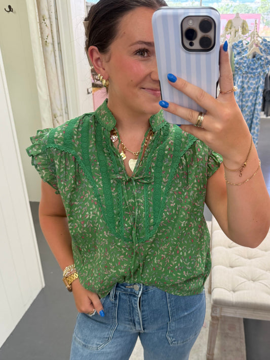Person wearing a green floral top and blue jeans taking a mirror selfie.